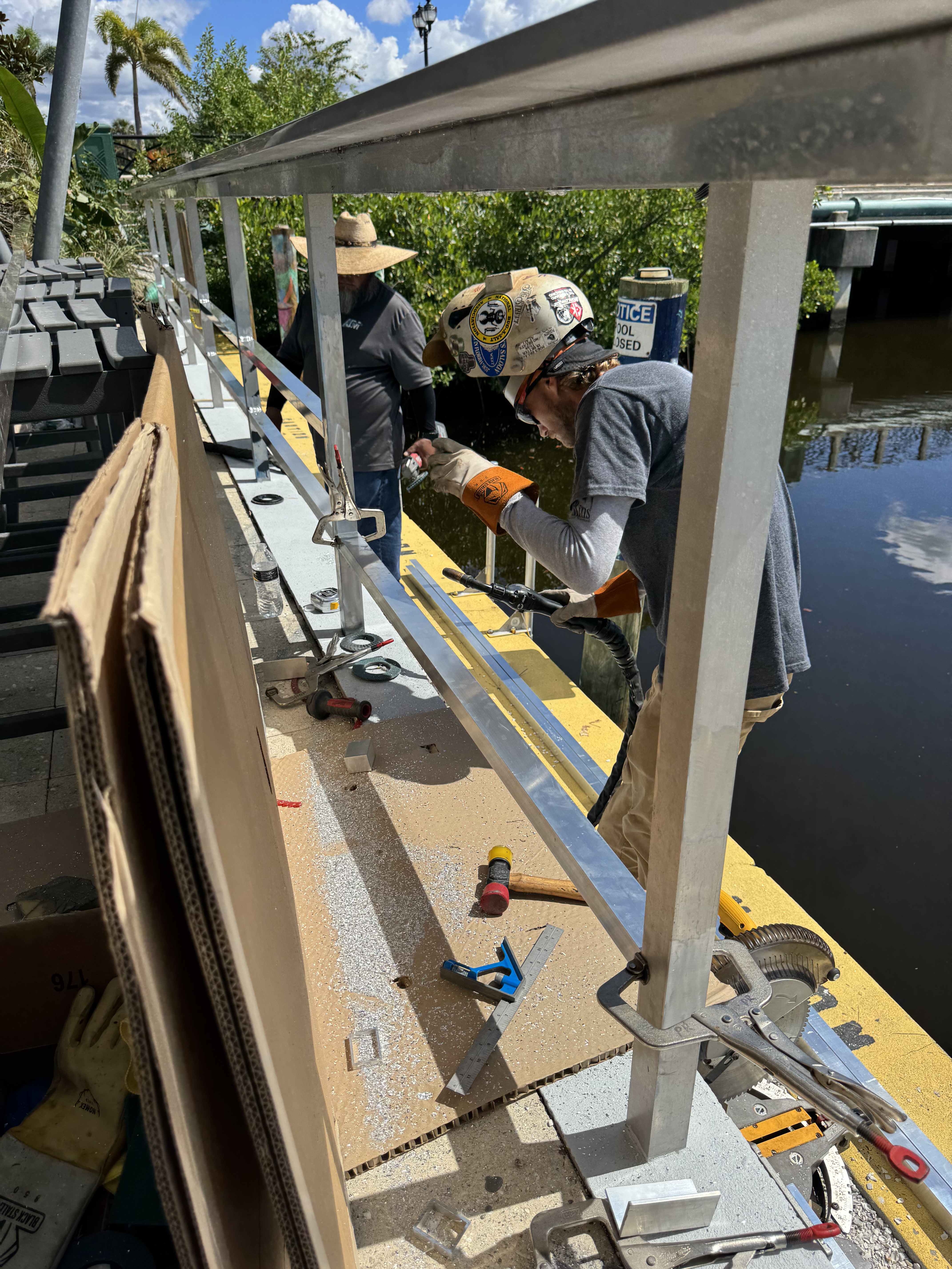 Metal fabrication work on a boat lift frame with workers assembling aluminum components at a waterfront dock.
